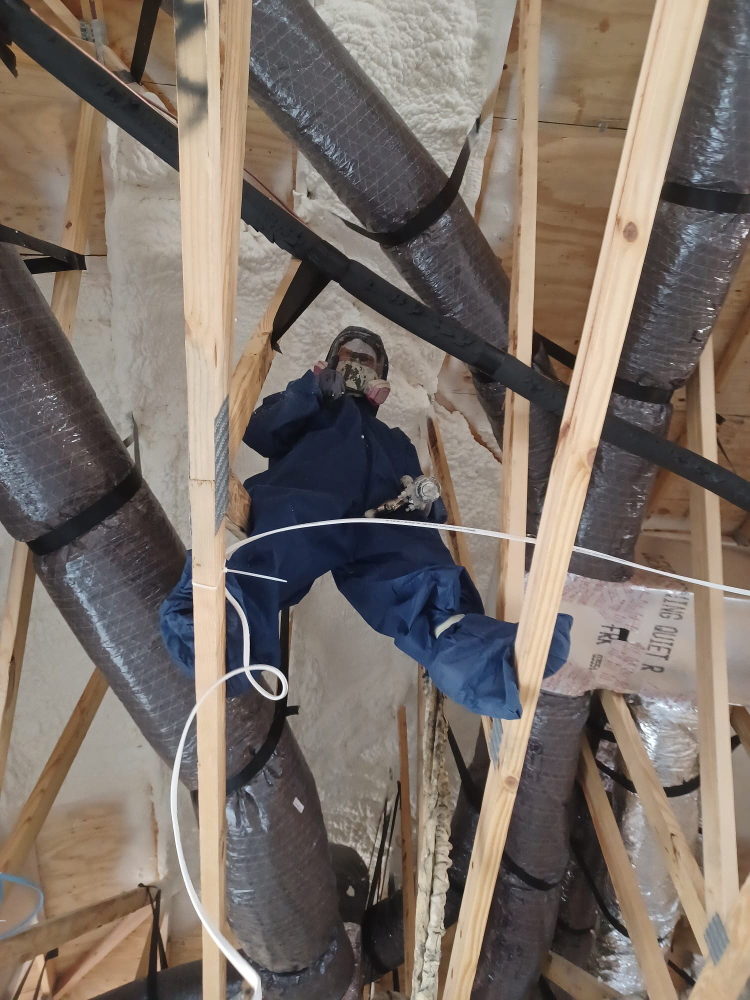 Spray foam installer working between attic trusses beside HVAC ducts